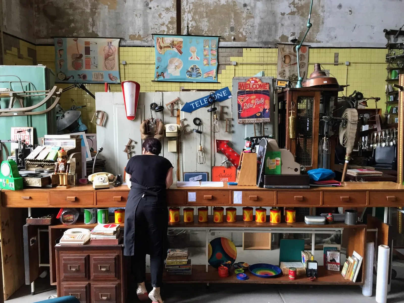 A person working at a long wooden counter in a reuse and repair workshop, the wall behind them hung with salvaged signs, lamps, telephones and anatomical charts.